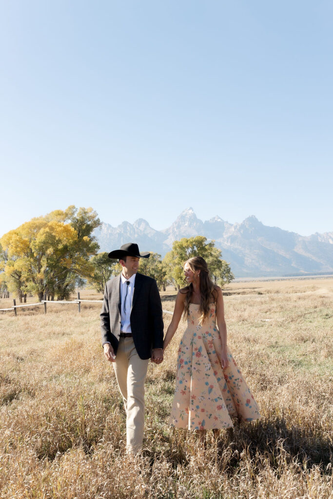 A man and woman holding hands walking in a grassy field with the grand Teton mountains behind them in Jackson Hole Wyoming